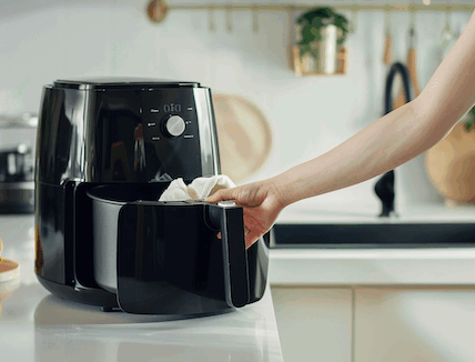Ein Airfryer steht auf einem weißen Küchencounter. Er ist offen und wird mit einem weissen Tuch von Innen ausgewischt. Den Airfryer reinigen leicht gemacht